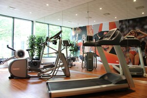 Un gimnasio con diversos equipos de ejercicio, amplias ventanas y un mural en la pared.
