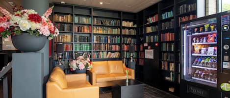 A modern library with a yellow sofa, bookshelves, a coffee machine, and a large flower arrangement.