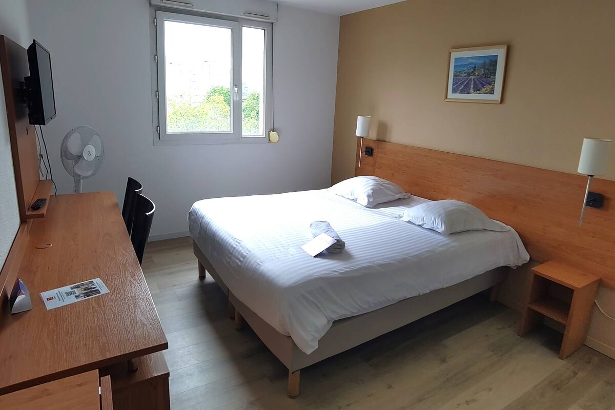 Hotelkamer met een bed, nachtkastjes, een bureau, een stoel, een televisie, een ventilator en een raam met uitzicht op groen.