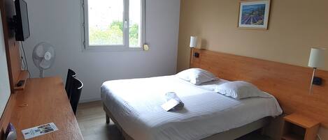A hotel room with a bed, bedside tables, a desk, a chair, a TV, a fan, and a window with a view of greenery.