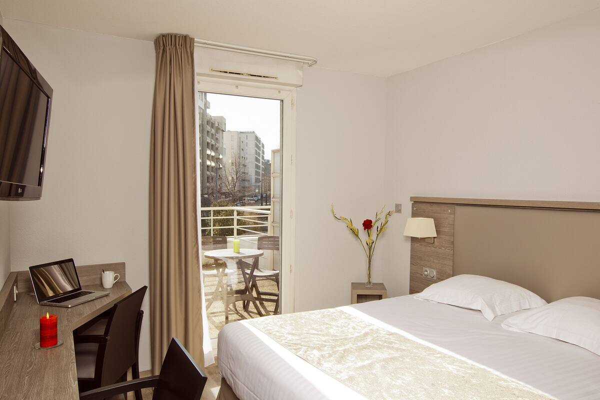 Een hotelkamer met een bed, een bureau met een laptop, een stoel, een klein tafeltje, een vaas met bloemen en een uitzicht op gebouwen door een raam.