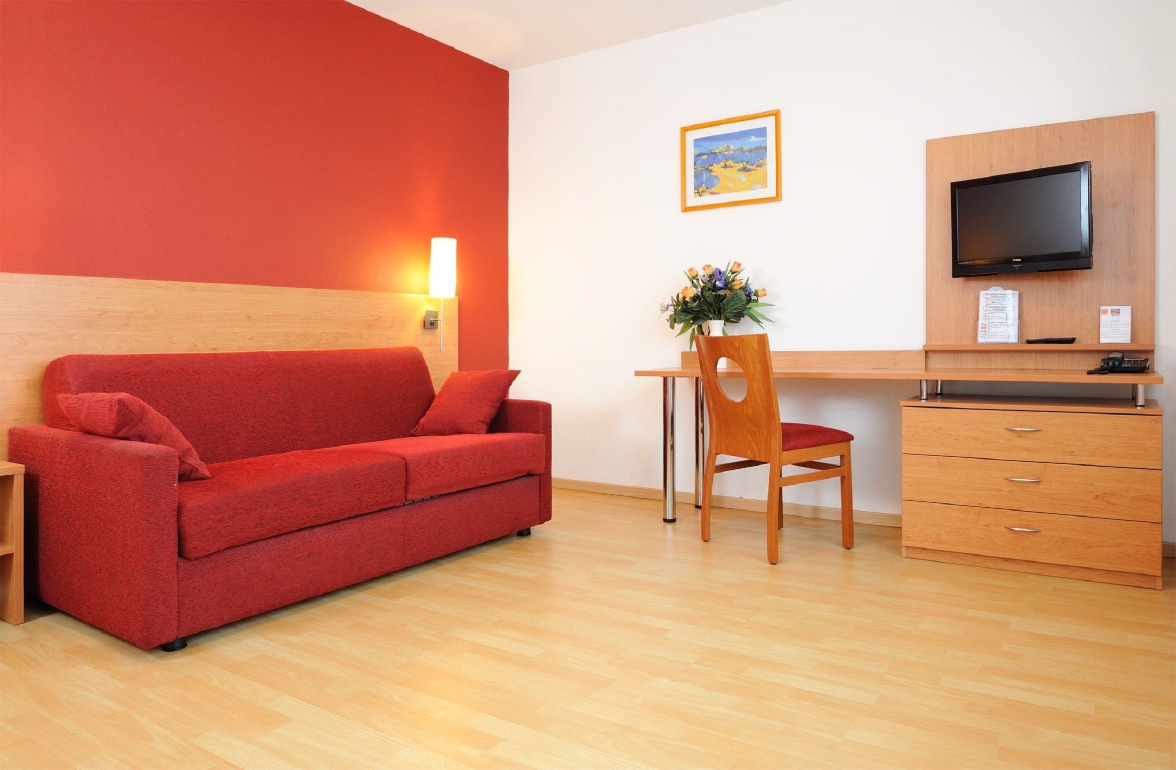 A room with a red sofa, a wooden desk, a chair, a TV, and a wooden cabinet.
