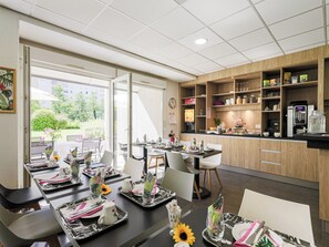 Un espace repas moderne dans un hôtel, avec des tables préparées pour le petit-déjeuner, une machine à café et une vue sur l’extérieur.
