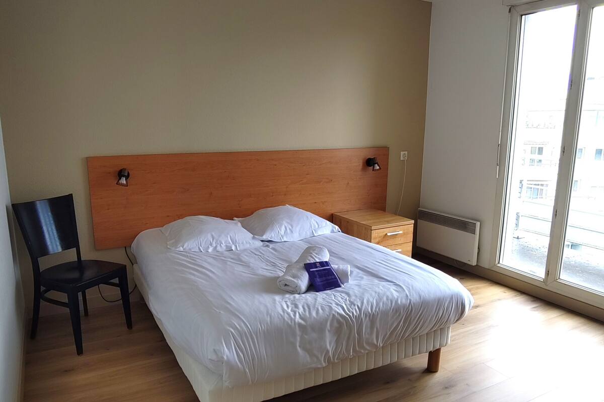 Een net opgemaakt bed met witte lakens, een houten hoofdbord en een nachtkastje met een lamp.