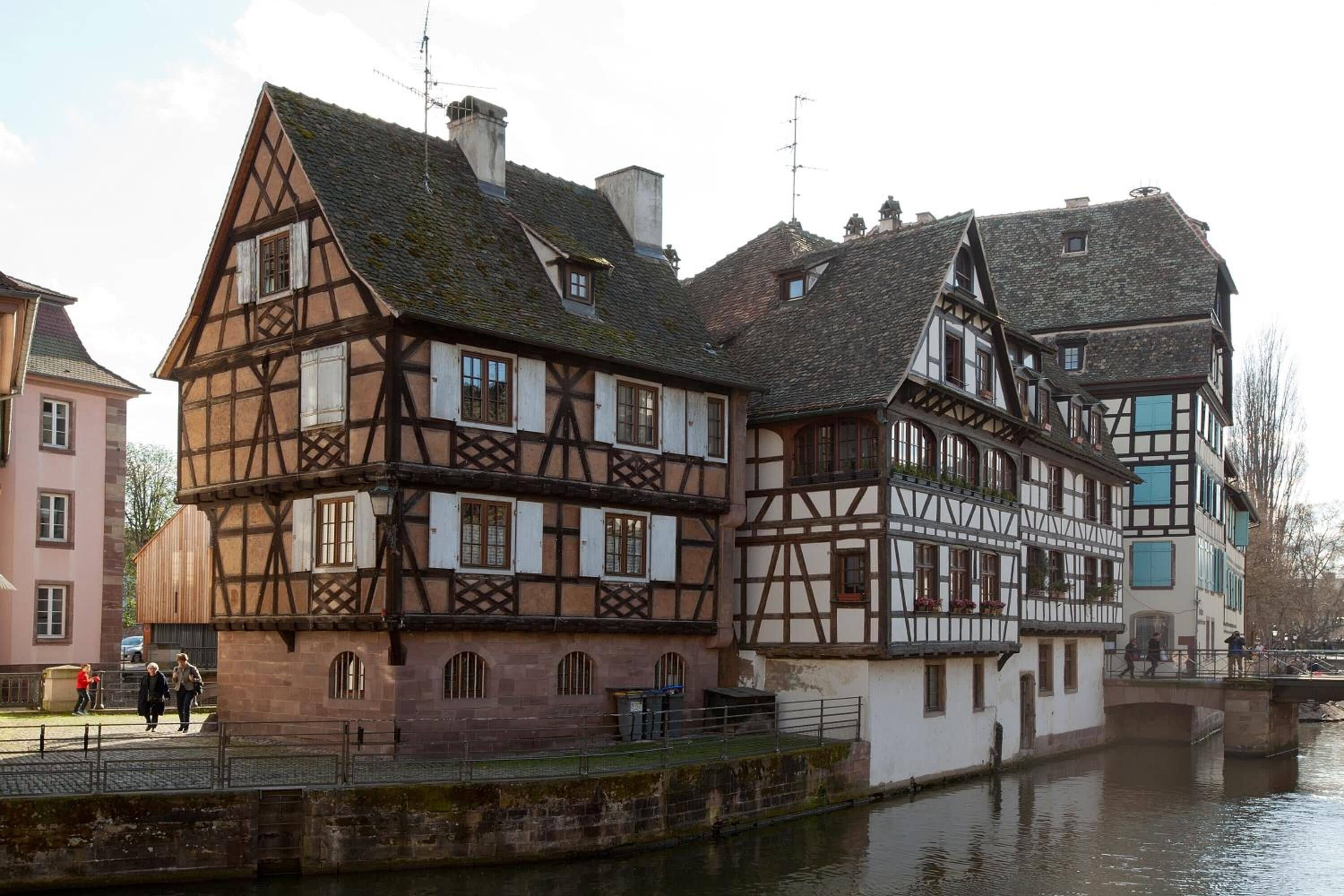 A traditional half-timbered building with a river in front, featuring a bridge and a pedestrian walkway.
