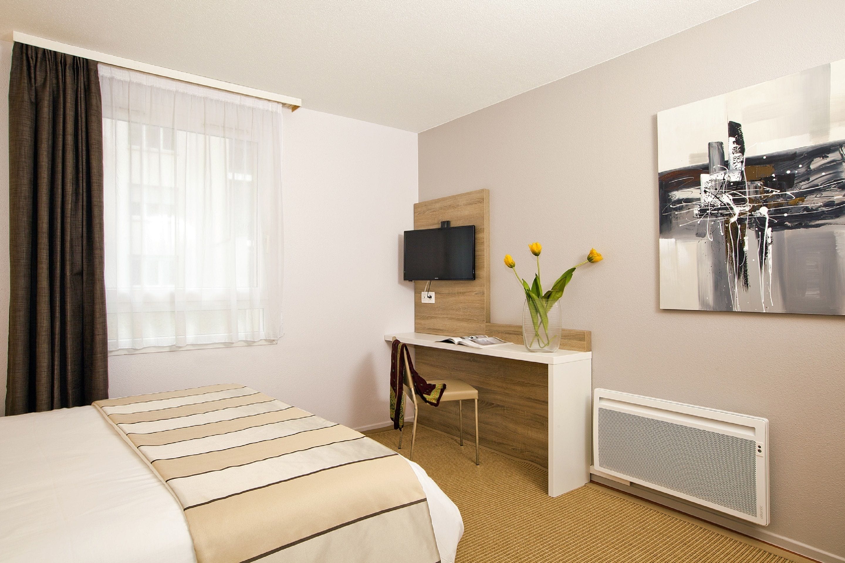 Hotelkamer met een bed, bureau, stoel, televisie, vaas met bloemen en een radiator.
