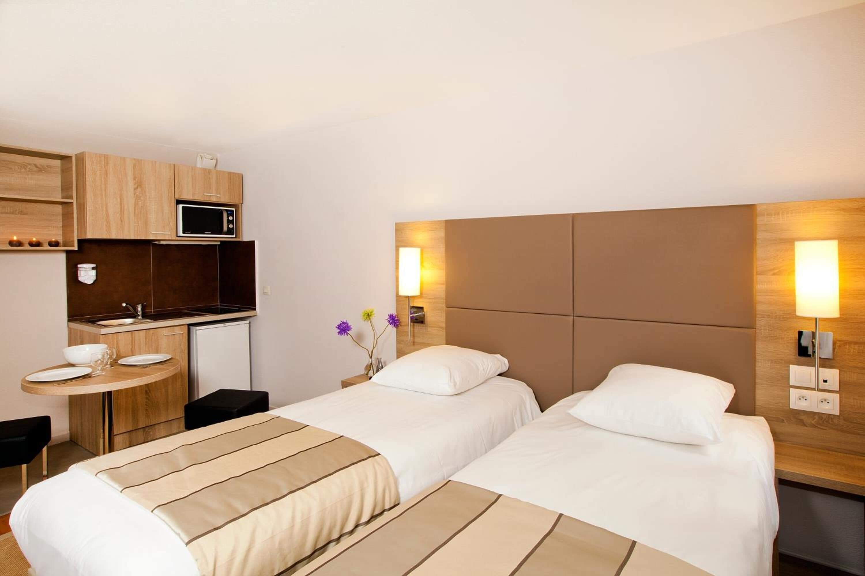 Une chambre d’hôtel avec deux lits, une petite kitchenette, une table à manger ronde et une table de chevet avec une lampe.