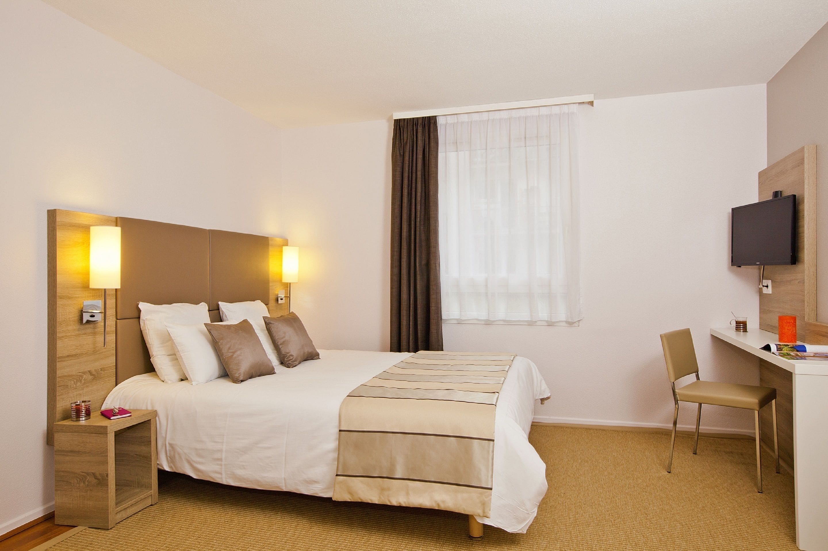 Una habitación de hotel con cama, mesita de noche, silla, escritorio, televisor y ventana con cortinas.
