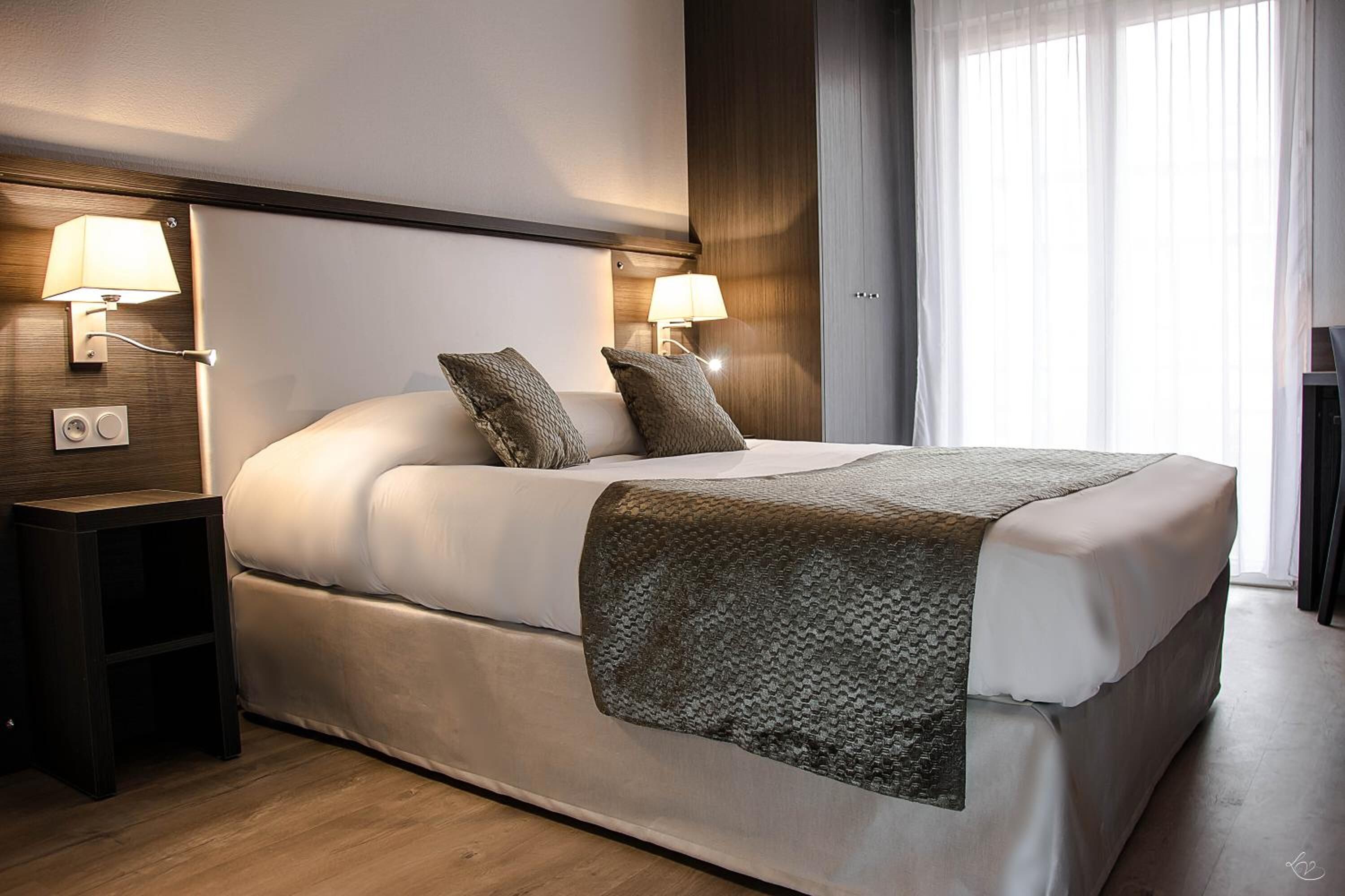 Una habitación de hotel moderna con una cama grande, dos lámparas de noche, una mesita de noche de madera y un gran ventanal con cortinas translúcidas.