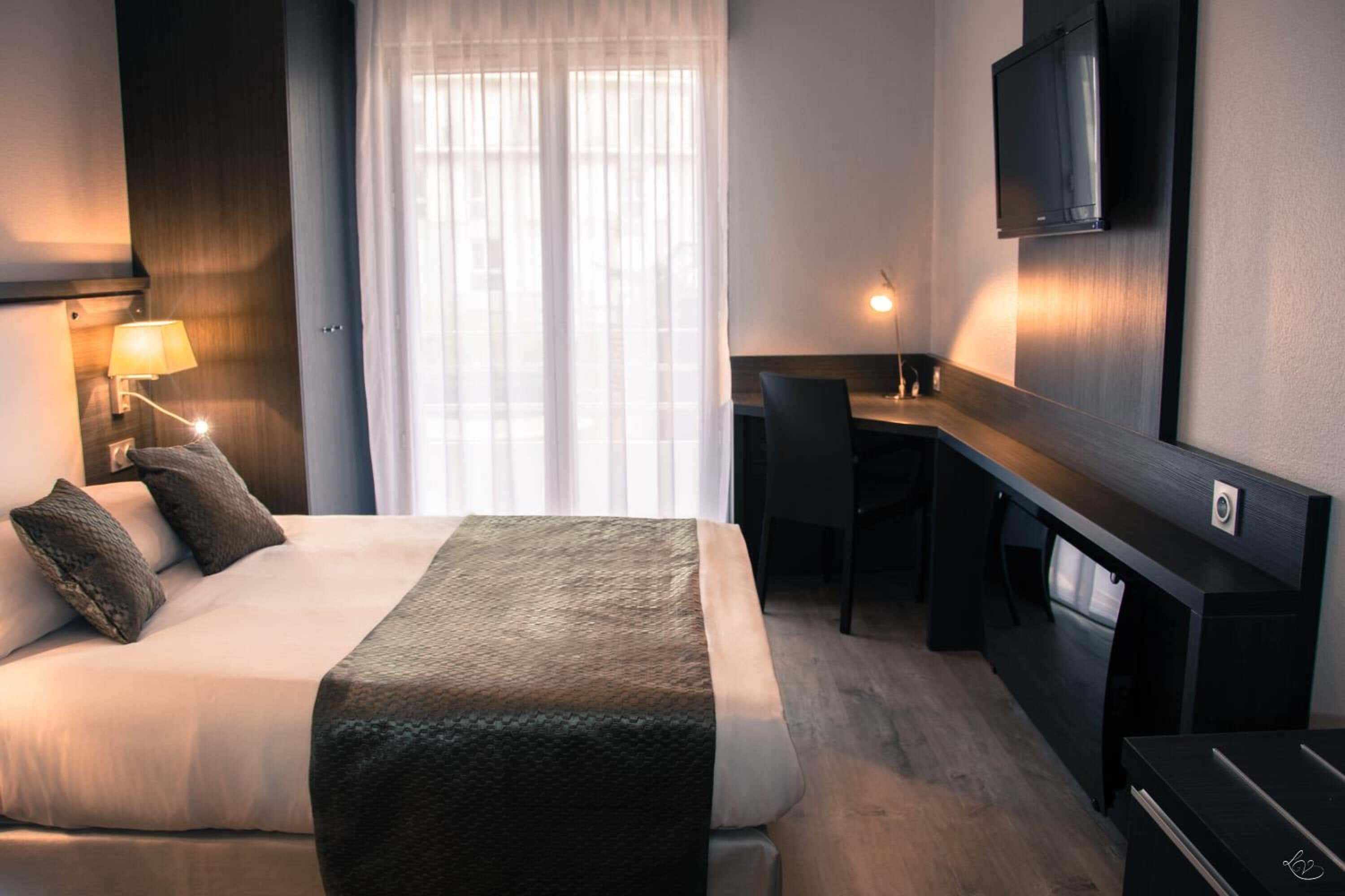 Habitación de hotel con cama, escritorio, silla, televisor y ventana con cortinas.