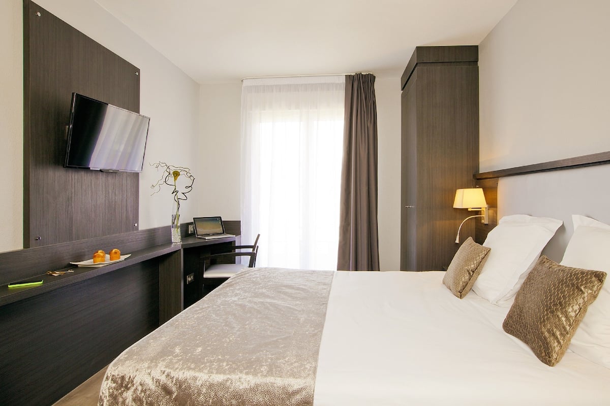 A hotel room with a bed, a desk with a laptop, a TV mounted on the wall, and a window with curtains.
