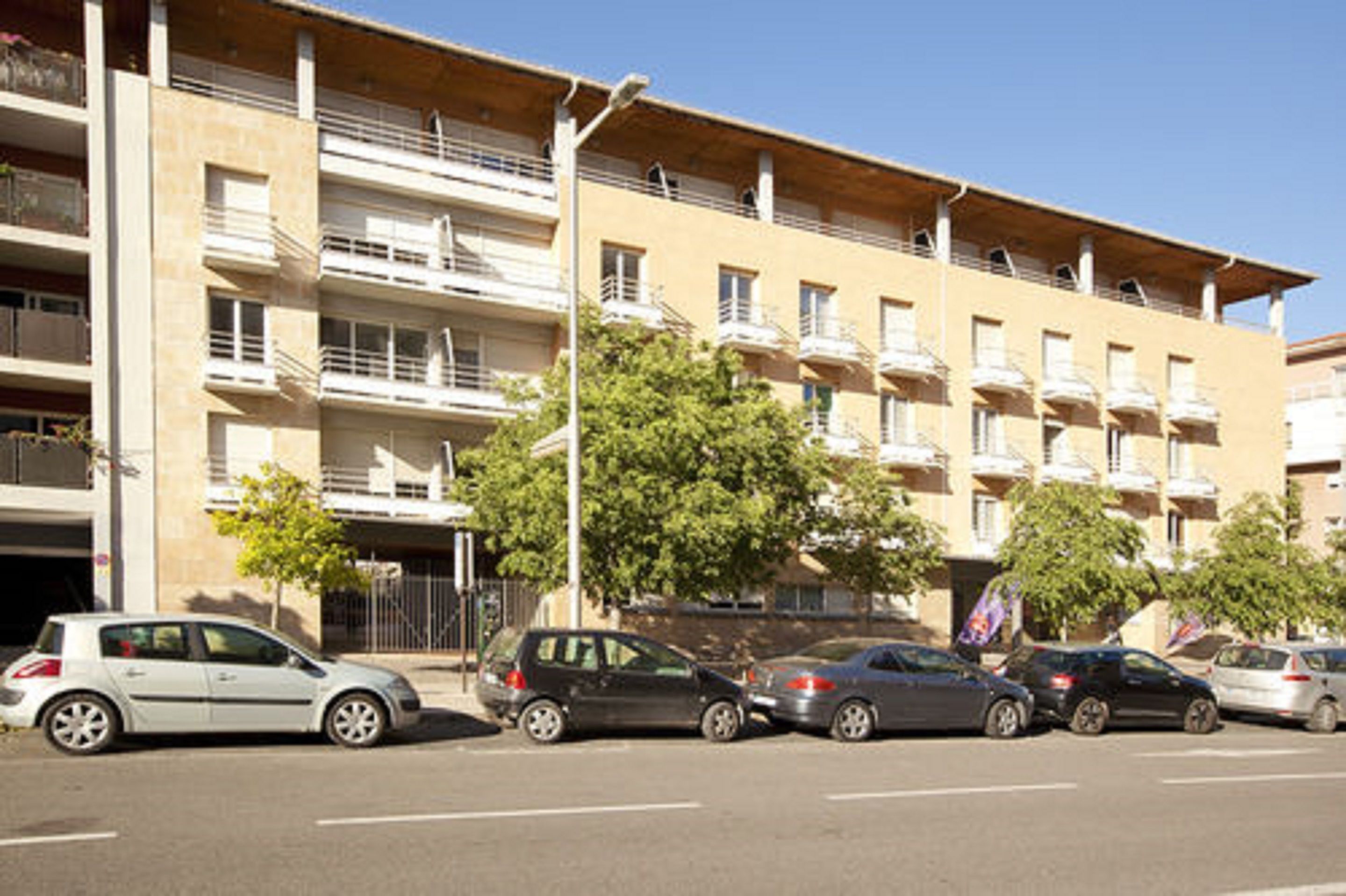 Un edificio residencial de varios pisos con balcones, coches aparcados y cielo despejado.