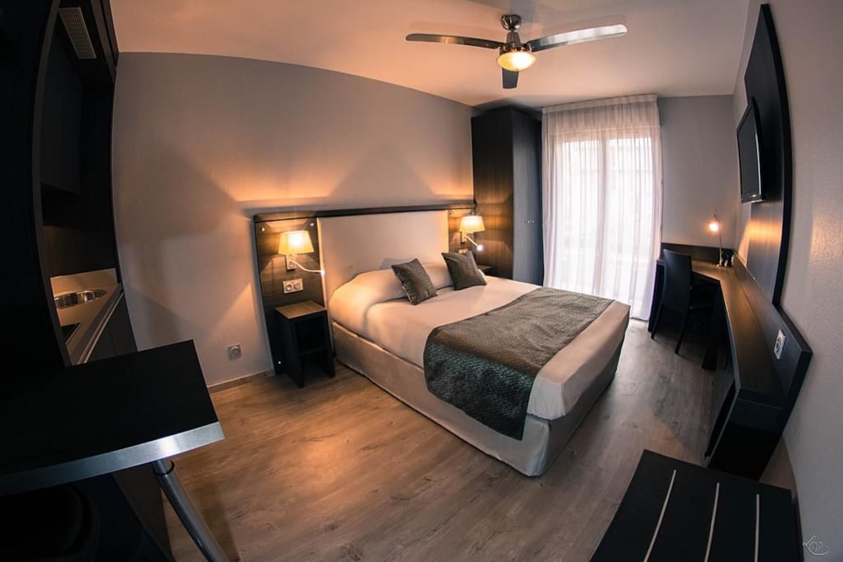 Habitación de hotel moderna con cama, lámparas de noche, ventilador de techo, televisor y un gran ventanal con cortinas.