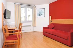 A room with a red sofa, wooden chairs, a desk with a laptop, a television, and a window with curtains.