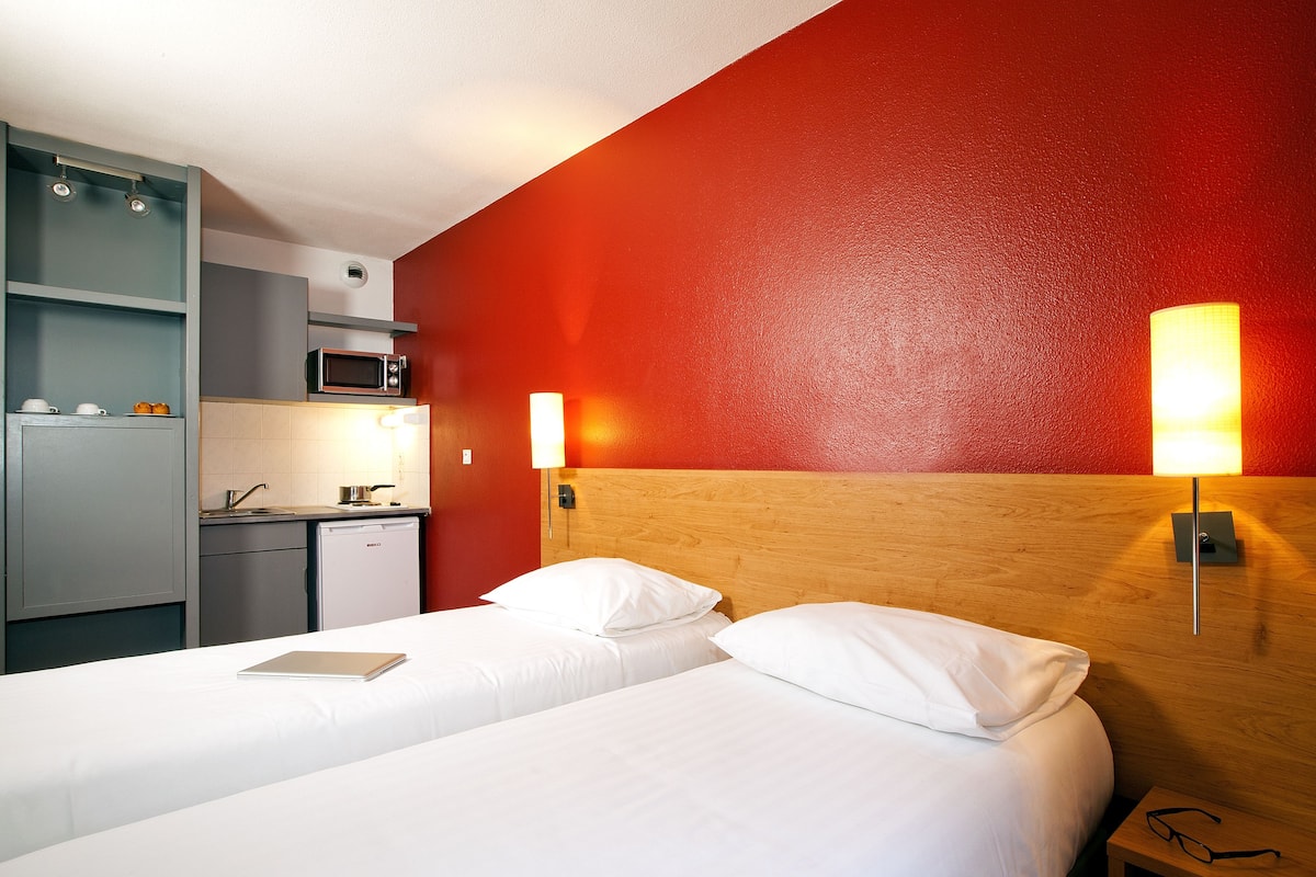 Une chambre d’hôtel comprenant un lit, des lampes de chevet, une table de chevet avec des verres, un petit coin cuisine équipé d’un four à micro-ondes et un mur d’accent de couleur rouge.