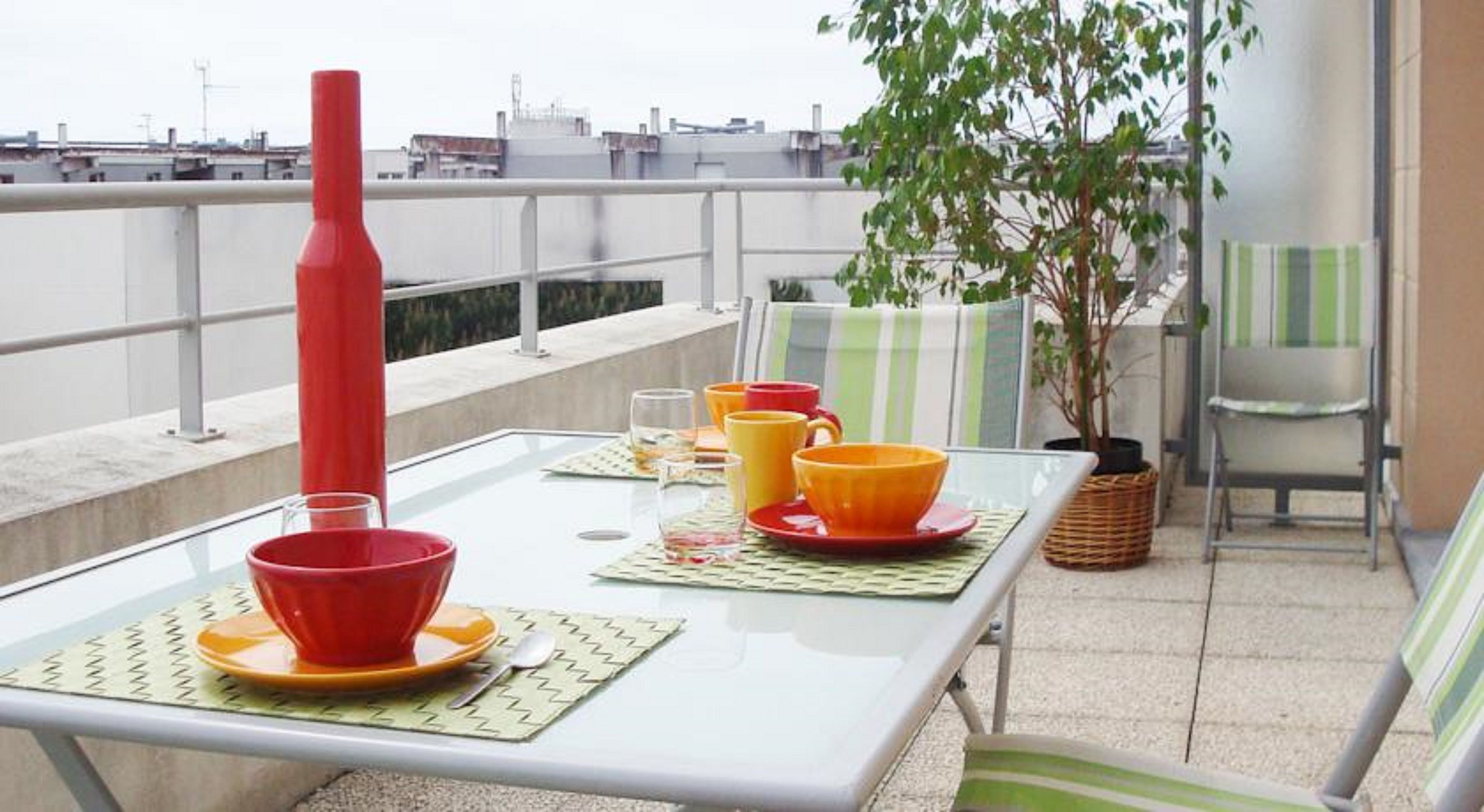 A balcony with a table set for two, featuring a red vase, orange and yellow bowls, and a potted plant.