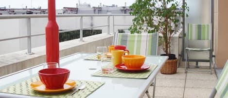 Ein Balkon mit Tisch für zwei Personen, mit einer roten Vase, orangefarbenen und gelben Schalen sowie einem Blumentopf.