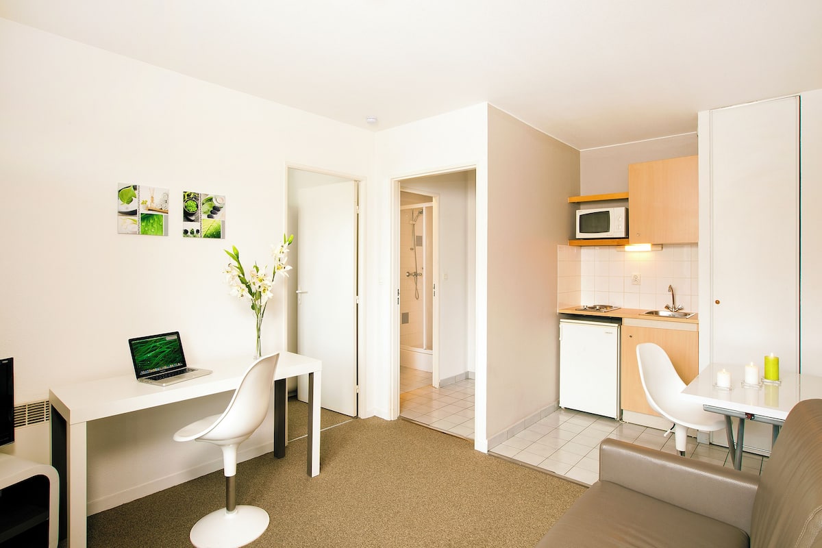 Une pièce moderne et bien éclairée, équipée d’un bureau, de deux chaises, d’une petite table avec des bougies, d’un four à micro-ondes et d’une petite cuisine compacte.