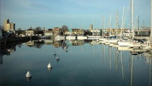 En lysthavn med flere sejlbåde i havnen, roligt vand og bygninger i baggrunden.
