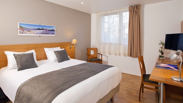 A hotel room with a large bed, a desk with a computer, a chair, a lamp, and a window with curtains.