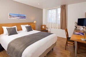 A hotel room with a large bed, a desk with a computer, a chair, a lamp, and a window with curtains.