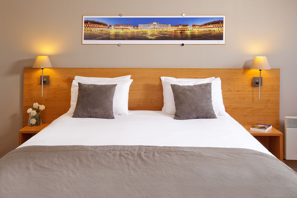 Ein Hotelzimmer mit einem großen Bett, zwei Nachttischlampen, einem Nachttisch mit einem Buch und einem Rahmenbild einer Stadtansicht an der Wand.