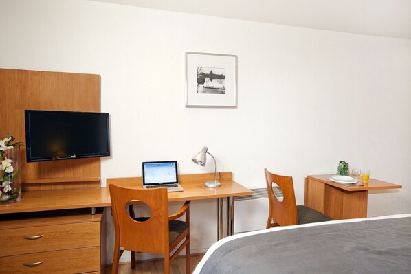 A hotel room with a wooden desk, a laptop, a desk lamp, a bed, a TV, and a framed picture on the wall.