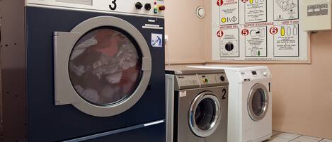 A laundry area with a large industrial washing machine, a smaller front-loading machine, and a wall-mounted instruction poster.