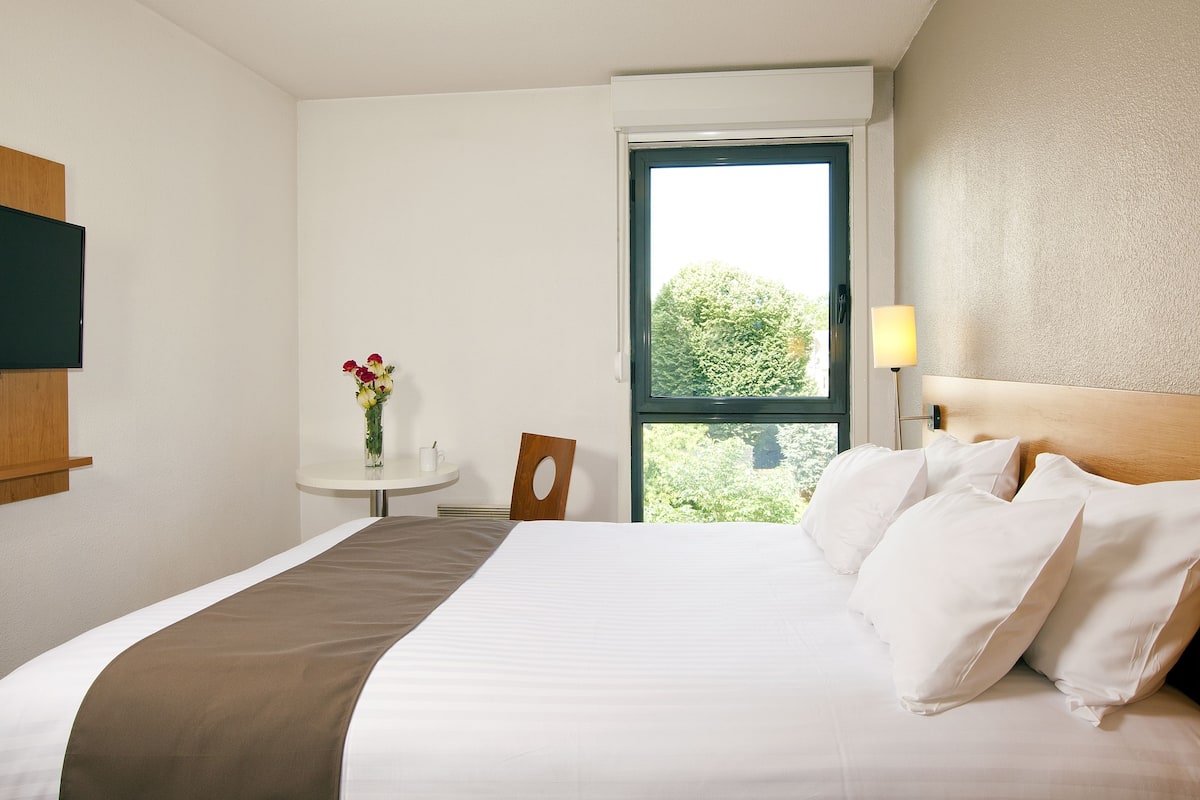 A hotel room with a bed, a small table with a vase of flowers, a chair, and a window with a view of trees.