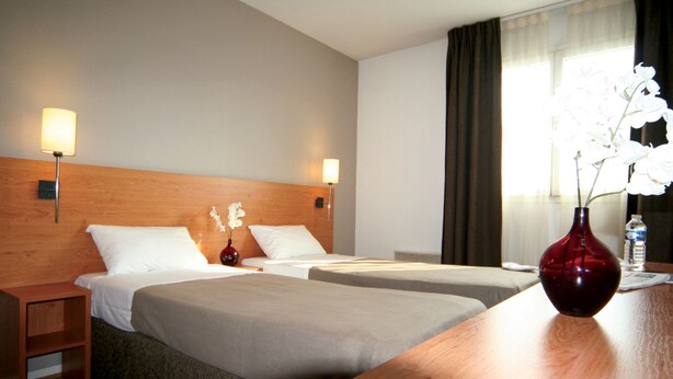 Habitación de hotel con dos camas, cabecero de madera, lámparas de noche, un jarrón con flores y una botella de agua sobre la mesita.
