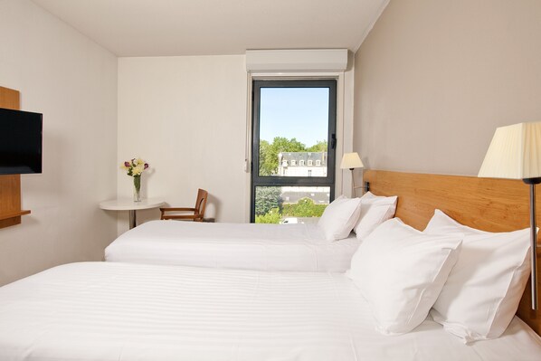 A hotel room with two beds, a small table with a vase of flowers, a chair, a TV, and a window with a view of buildings and trees.