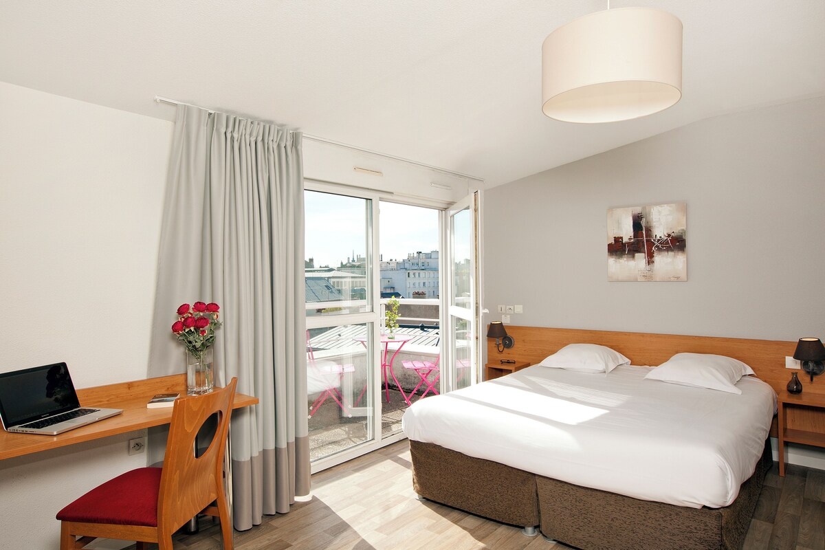 A hotel room with a bed, desk, laptop, chair, and a view through a glass door.