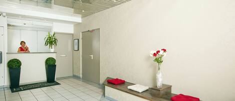 A reception area with a receptionist, two potted plants, a reception desk with red and white cushions, and a vase with flowers.