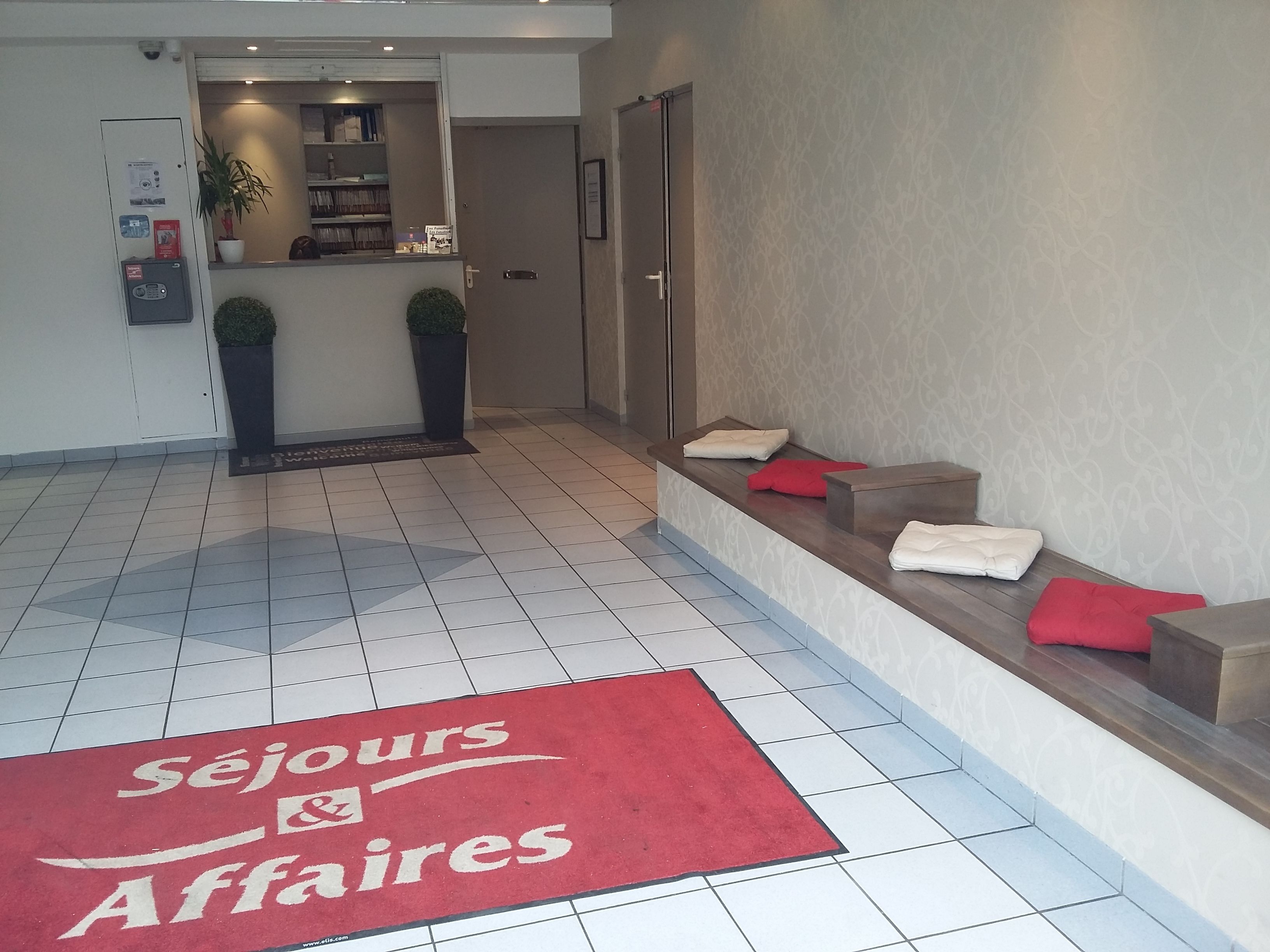 Eine Hotelhalle mit einer Rezeption, einem roten Teppich mit dem Schriftzug 'Sejours & Affaires' und einer Sitzecke mit Kissen.