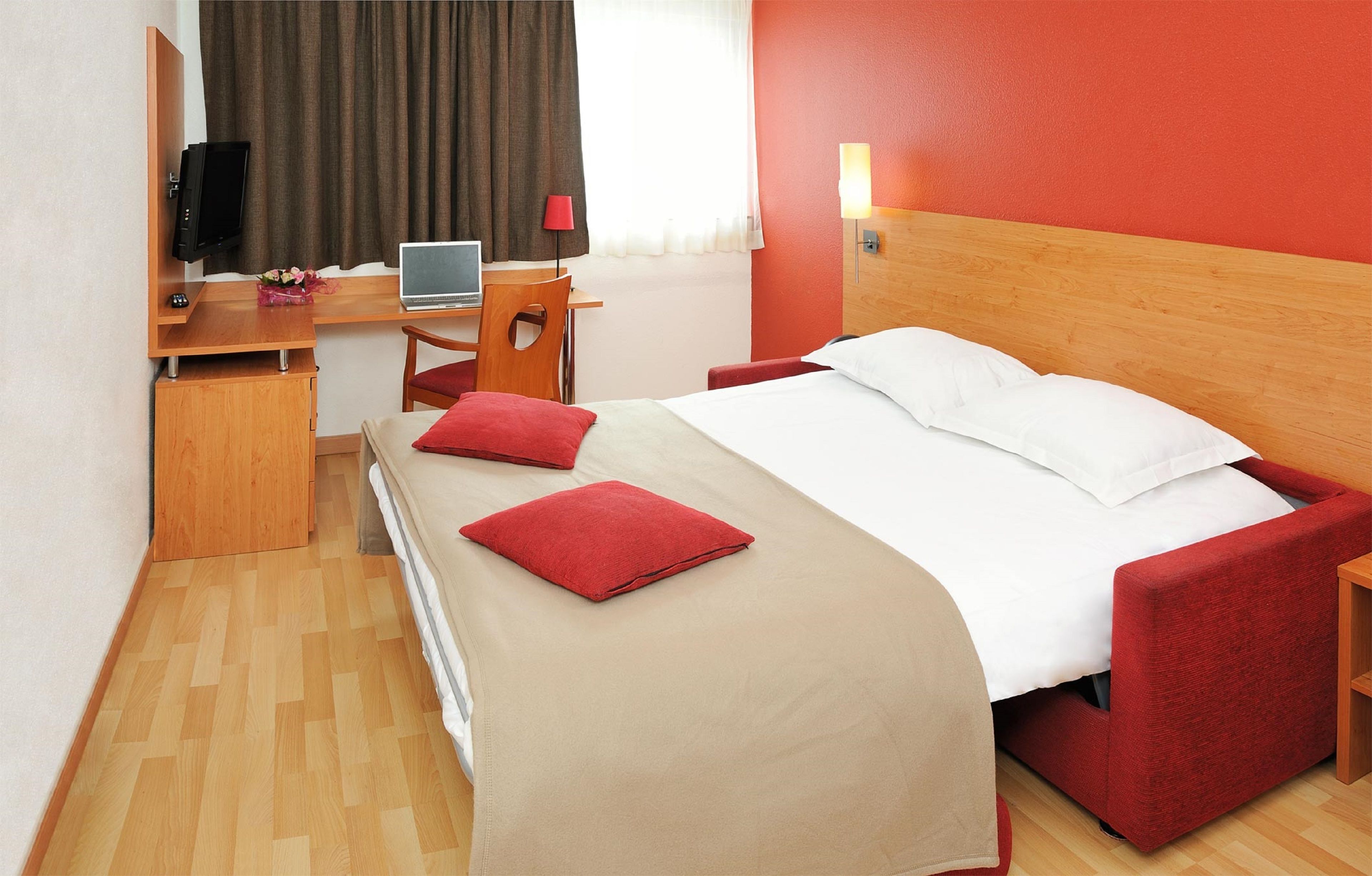 A hotel room with a bed, a desk with a laptop, a television, and a red chair.