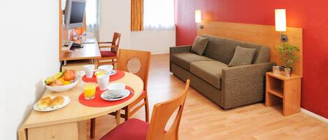 A hotel room with a dining area, a sofa, a TV, and a bedside table with a lamp.