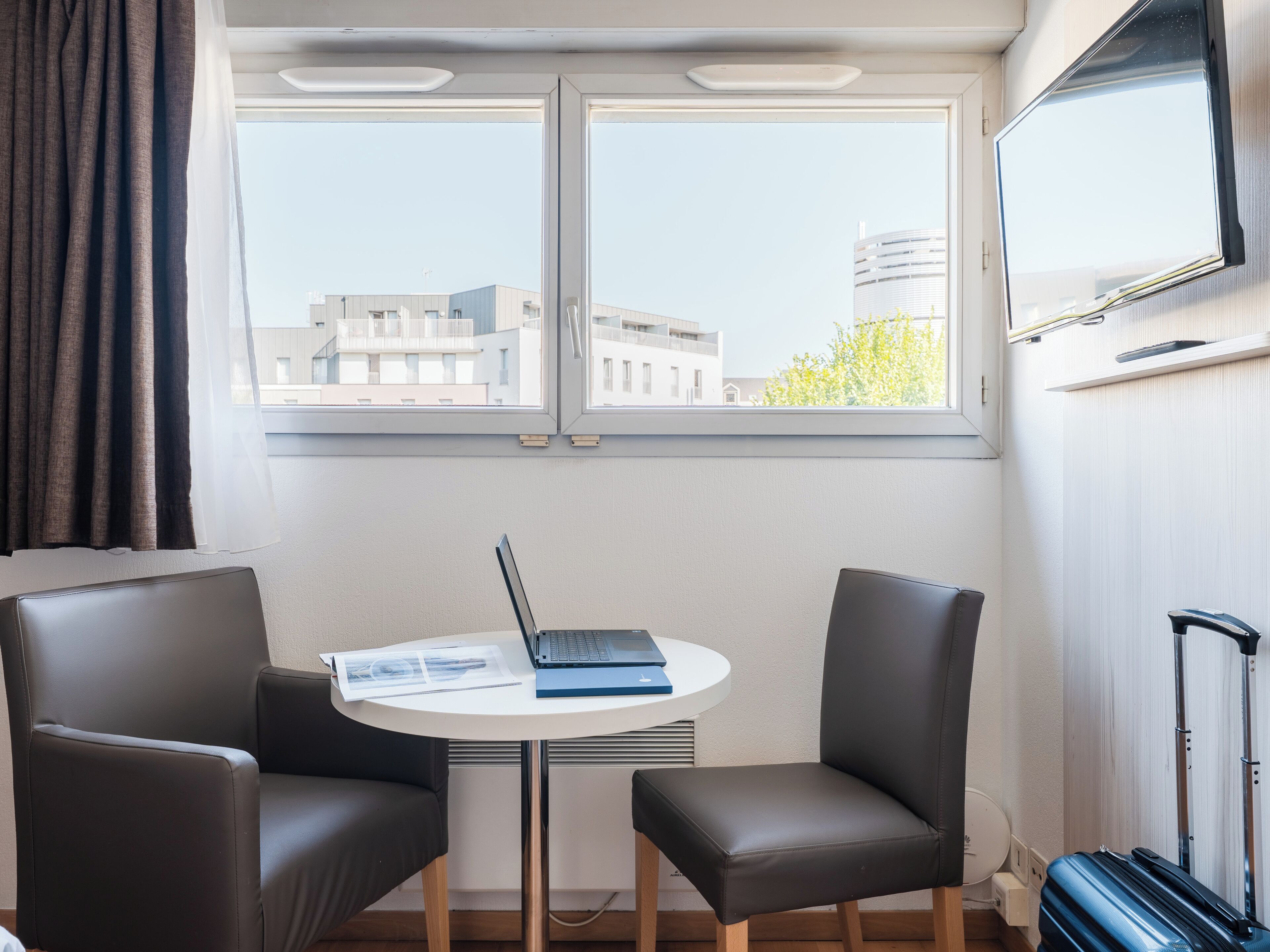 A room with a table, two chairs, a laptop, a suitcase, and a TV on the wall.