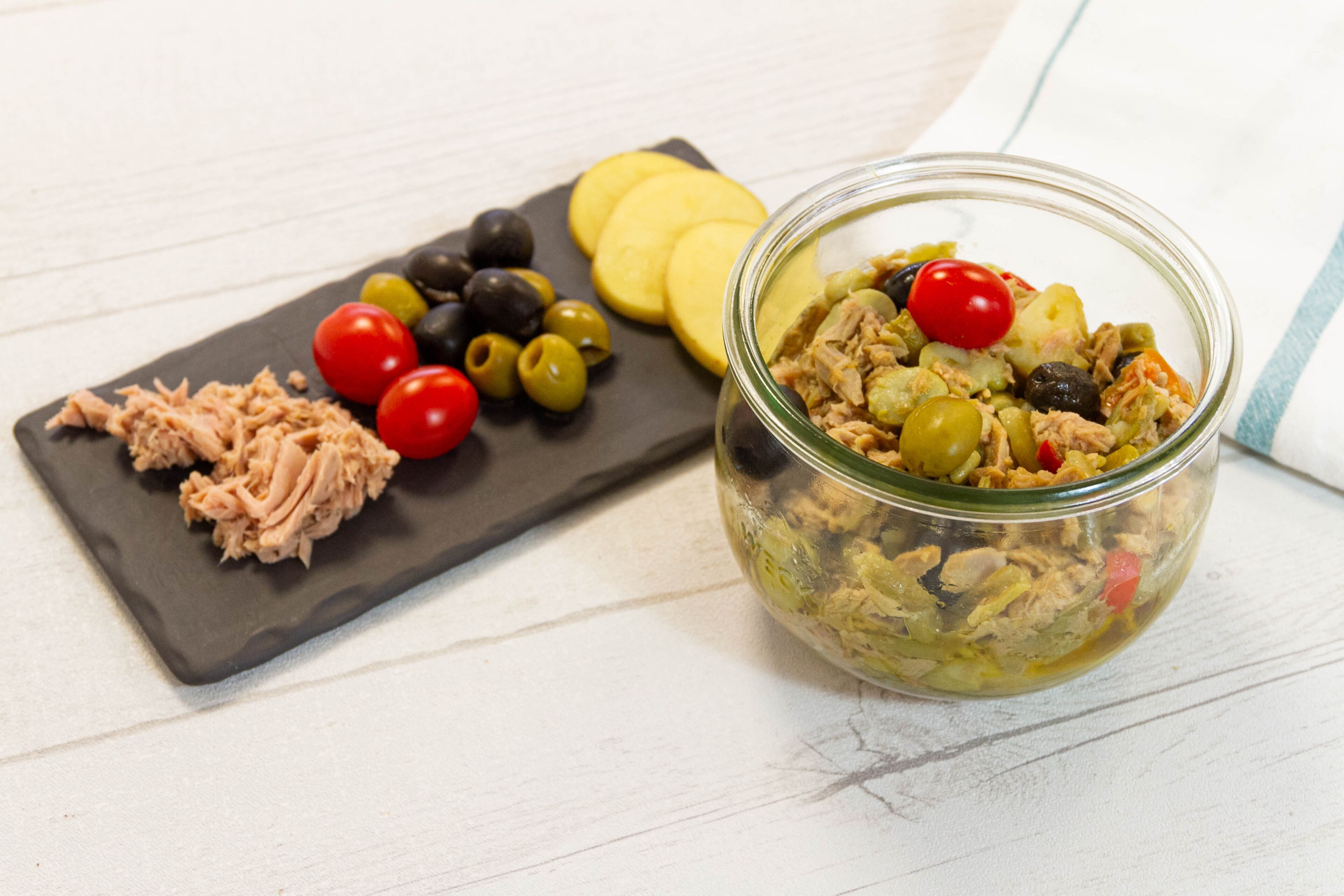 A glass jar filled with a salad containing tuna, olives, and cherry tomatoes, alongside a black slate board with sliced potatoes, olives, and cherry tomatoes.