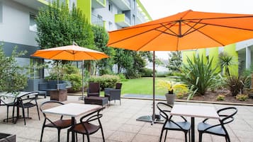 Ein Sitzbereich im Freien mit Tischen und Stühlen, zwei orangefarbene Sonnenschirme und ein Gebäude mit grün-weißen Außenwänden.