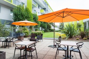 An outdoor seating area with tables and chairs, two orange umbrellas, and a building with green and white exterior walls. - Comfort Aparthotel Saint-Nazaire Les Portes de l Ocean (Saint-Nazaire)