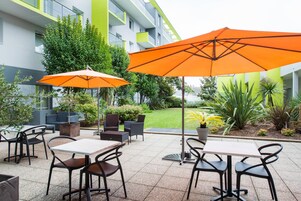 Un área de estar al aire libre con mesas y sillas, dos sombrillas naranjas y un edificio con paredes exteriores verdes y blancas.