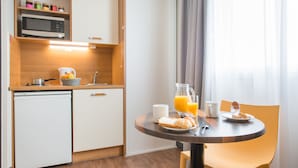 A compact kitchen with a microwave, sink, and a small refrigerator. There is a dining table with breakfast items like bread, a jug of orange juice, and a glass of water. - Comfort Aparthotel Saint-Nazaire Les Portes de l Ocean (Saint-Nazaire)