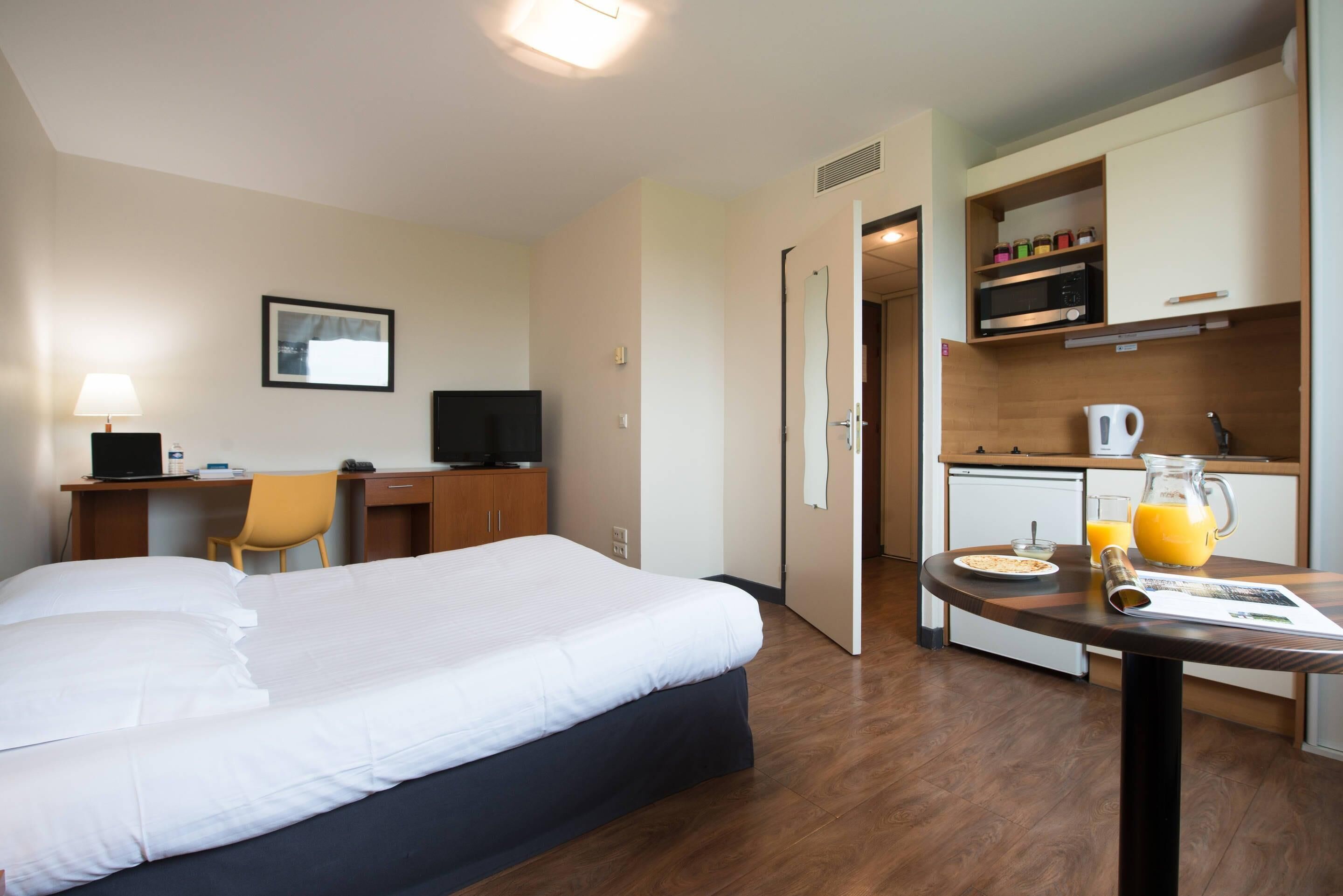 A hotel room with a bed, desk, chair, kitchenette, and breakfast table with a pitcher of orange juice.