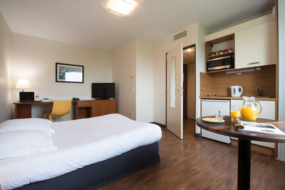 A hotel room with a bed, desk, chair, kitchenette, and breakfast table with a pitcher of orange juice.