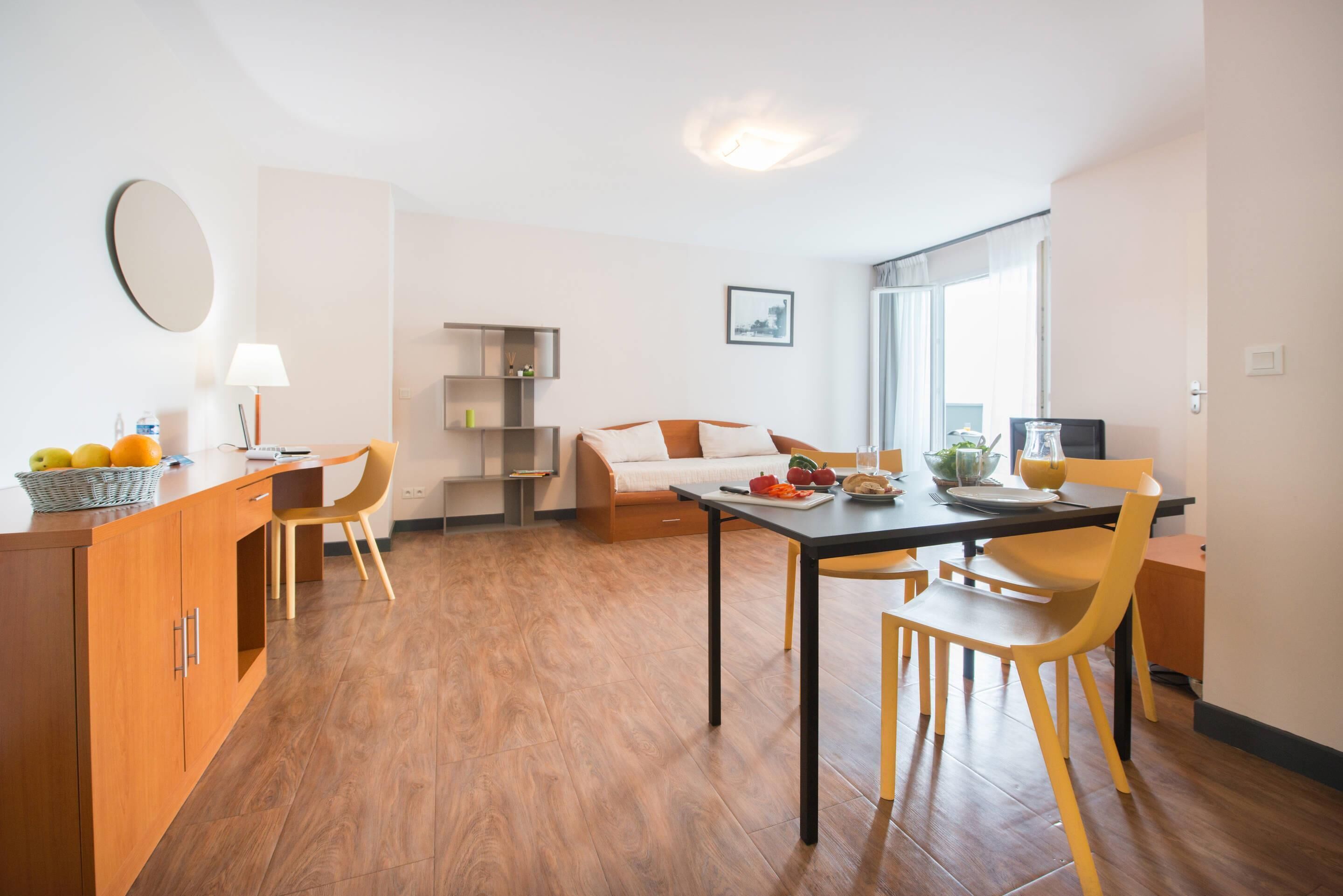 A modern kitchen with wooden cabinets, a dining table, and chairs. There is a sofa and a small table with a lamp in the living area.