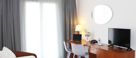 A hotel room with a bed, a desk with a laptop, a TV, a lamp, and a mirror.