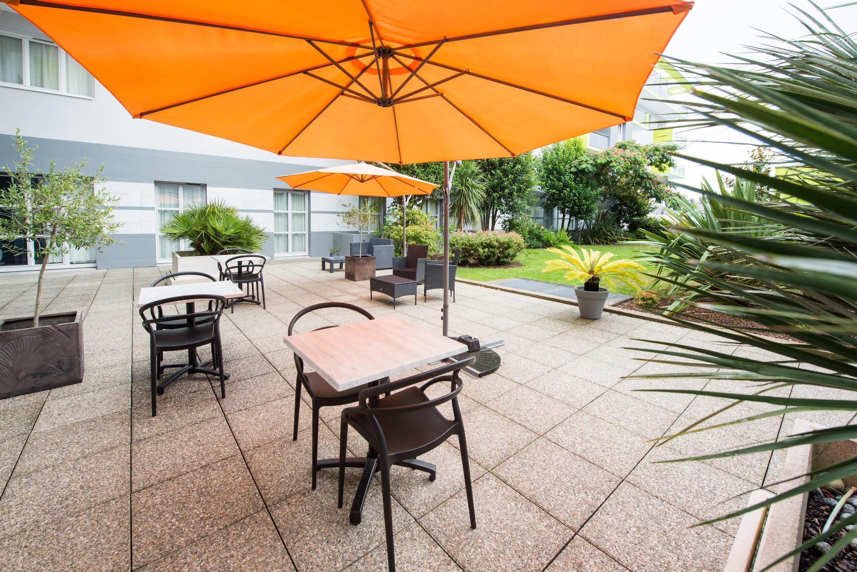 Un espace extérieur aménagé en terrasse, doté d’un sol carrelé, de plusieurs tables et chaises, et de parasols orange offrant de l’ombre.