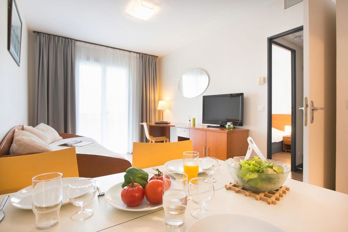 A hotel room with a dining area, a bed, a television, and a view of the outside.