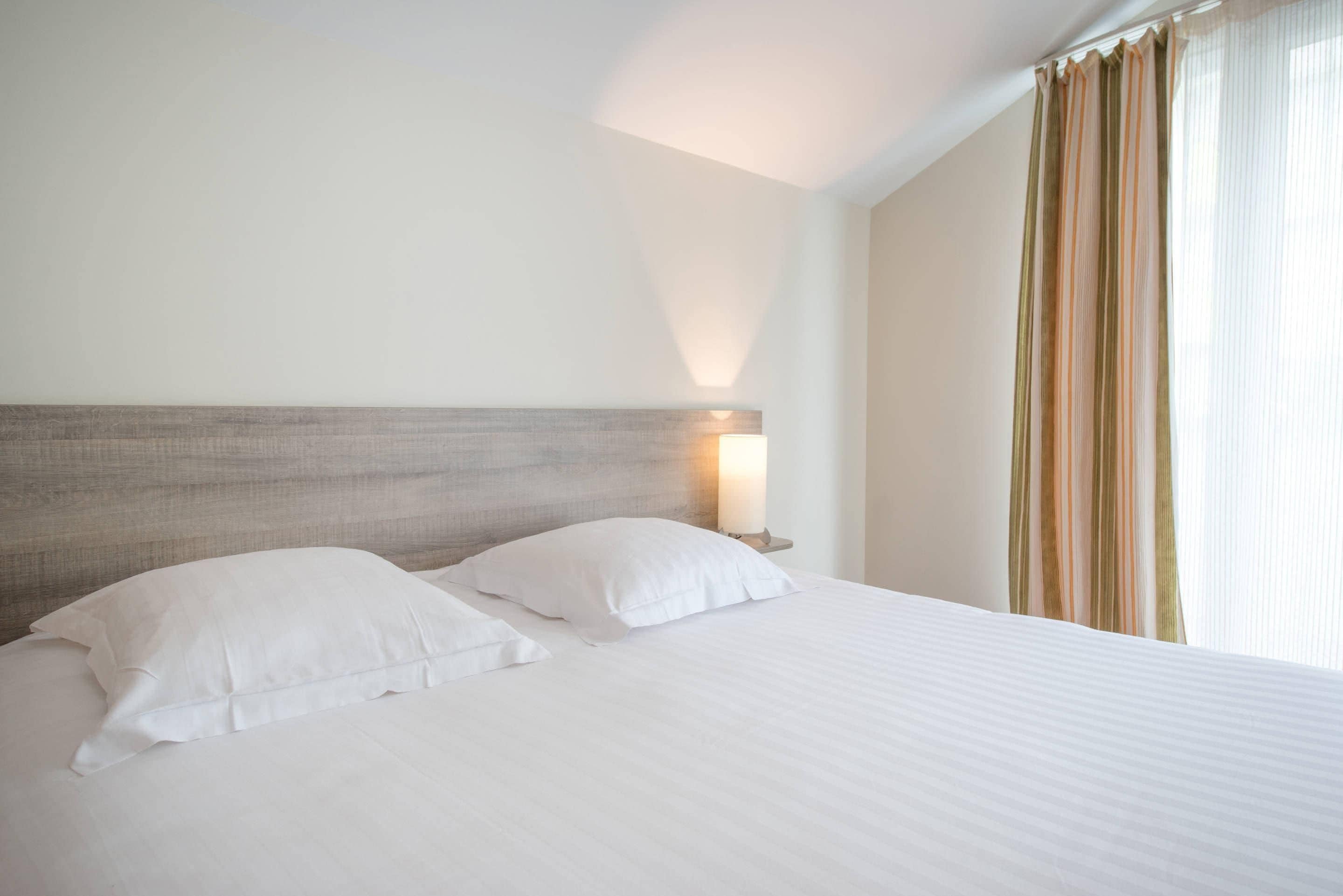 Habitación de hotel con cama, almohadas, cabecera, mesita de noche y ventana con cortinas.