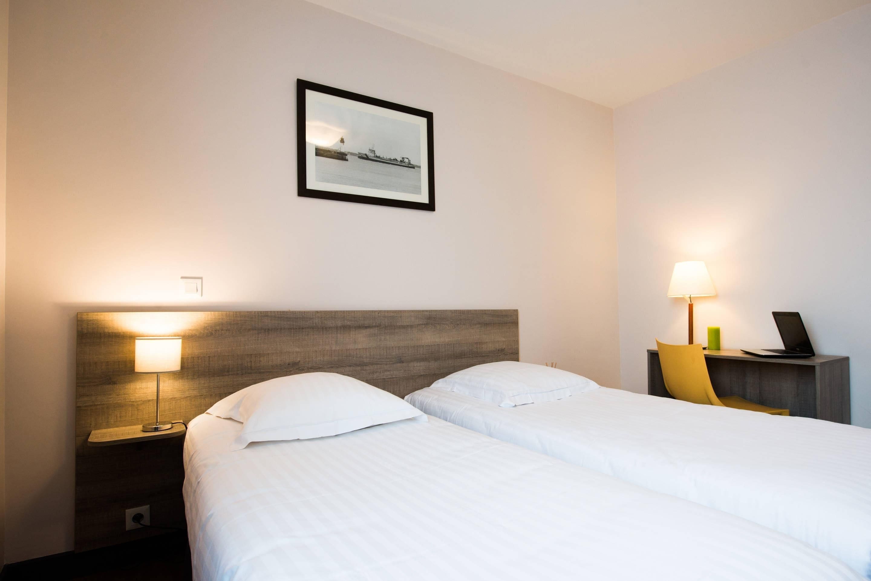 A hotel room with two beds, a nightstand with a lamp, a desk with a laptop, and a framed picture on the wall.